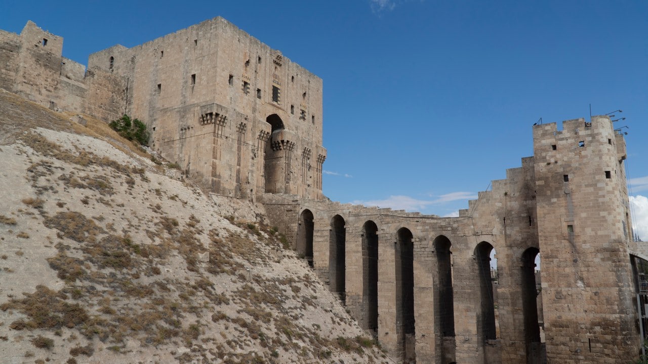 The Citadel of Aleppo in October 2023: a tall rectangular stone gate block on the crest of a steep stone-faced slope, with an arched gateway between projecting corbelled boxes, and a bridge on tall arched piers running to a square outer tower at the right, under a clear blue sky. The Citadel of Aleppo in October 2023: a tall rectangular stone gate block on the crest of a steep stone-faced slope, with an arched gateway between projecting corbelled boxes, and a bridge on tall arched piers running to a square outer tower at the right, under a clear blue sky.