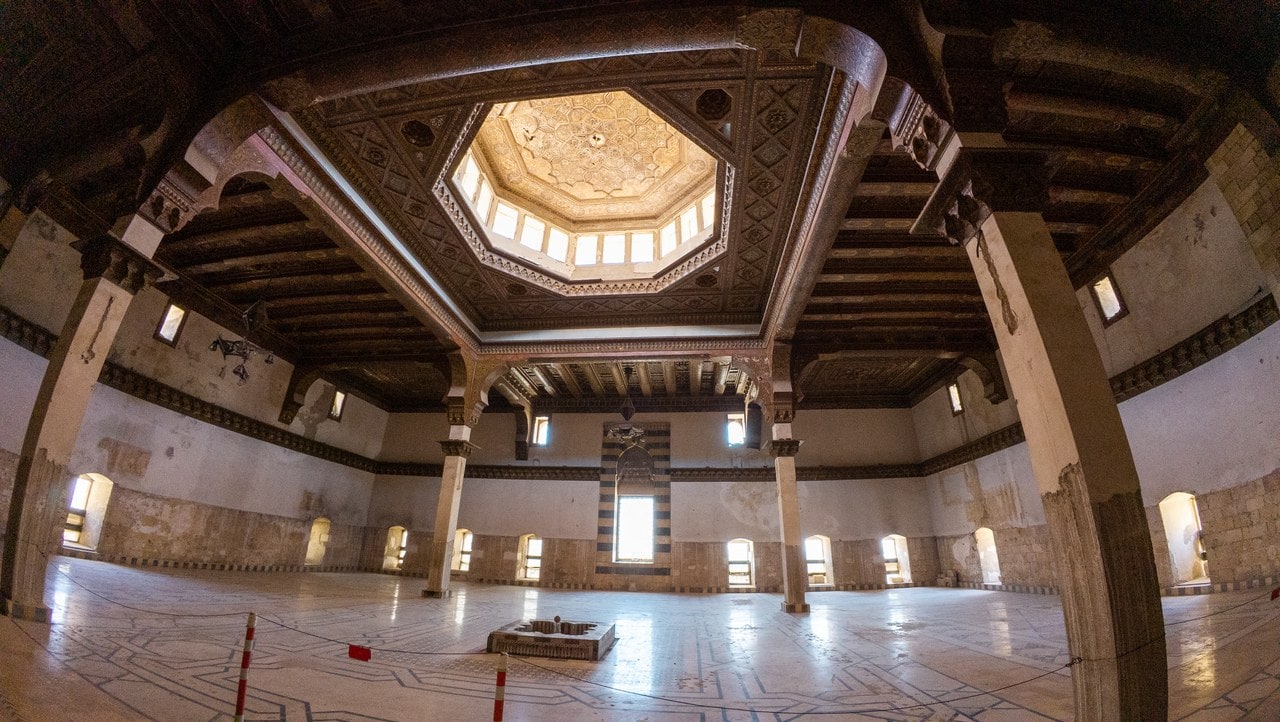 The interior of the throne hall in the Citadel of Aleppo in 2025, a large square room where four piers carry a carved and coffered wooden ceiling around a central octagonal lantern of small windows, over a marble floor inlaid with geometric bands, and a tall recess of striped black-and-white masonry in the far wall. The interior of the throne hall in the Citadel of Aleppo in 2025, a large square room where four piers carry a carved and coffered wooden ceiling around a central octagonal lantern of small windows, over a marble floor inlaid with geometric bands, and a tall recess of striped black-and-white masonry in the far wall.