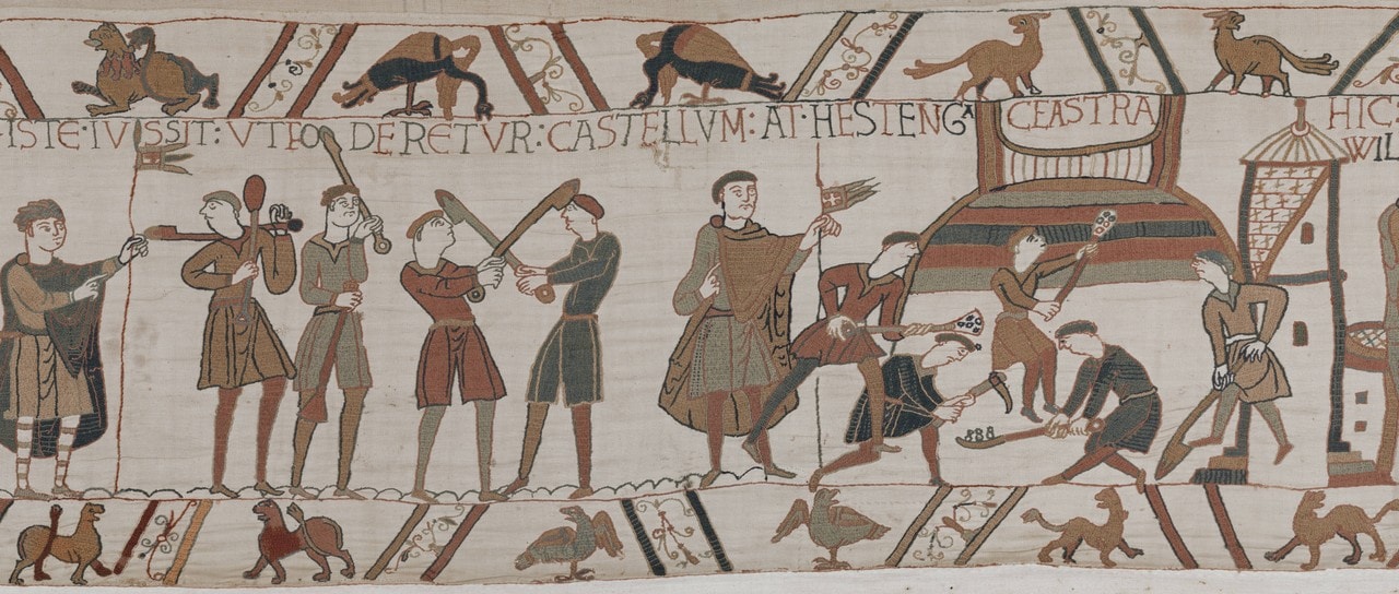 A panel of the Bayeux Tapestry beneath an inscription reading ISTE IUSSIT UT FODERETUR CASTELLUM AT HESTENGA CEASTRA: at the left men carry long-handled spades and mattocks, a figure in a green tunic stands holding a small pennant at the center, and at the right workmen heap up a mound of earth worked in colored bands. The next scene's inscription begins at the right edge of the frame. A panel of the Bayeux Tapestry beneath an inscription reading ISTE IUSSIT UT FODERETUR CASTELLUM AT HESTENGA CEASTRA: at the left men carry long-handled spades and mattocks, a figure in a green tunic stands holding a small pennant at the center, and at the right workmen heap up a mound of earth worked in colored bands. The next scene's inscription begins at the right edge of the frame.