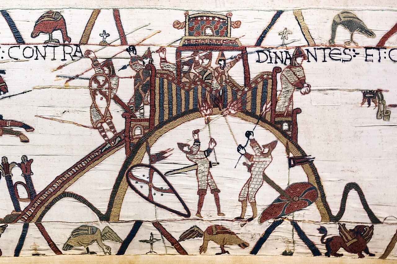 A detail of the Bayeux Tapestry: a small timber tower stands on the crown of a steep mound behind a palisade of vertical planks, defenders with spears above it, while two men in mail hold torches to the timbers at the foot and flames are stitched in red where the fire catches. Part of an inscription reading CONTRA DINANTES ET runs across the top. A detail of the Bayeux Tapestry: a small timber tower stands on the crown of a steep mound behind a palisade of vertical planks, defenders with spears above it, while two men in mail hold torches to the timbers at the foot and flames are stitched in red where the fire catches. Part of an inscription reading CONTRA DINANTES ET runs across the top.