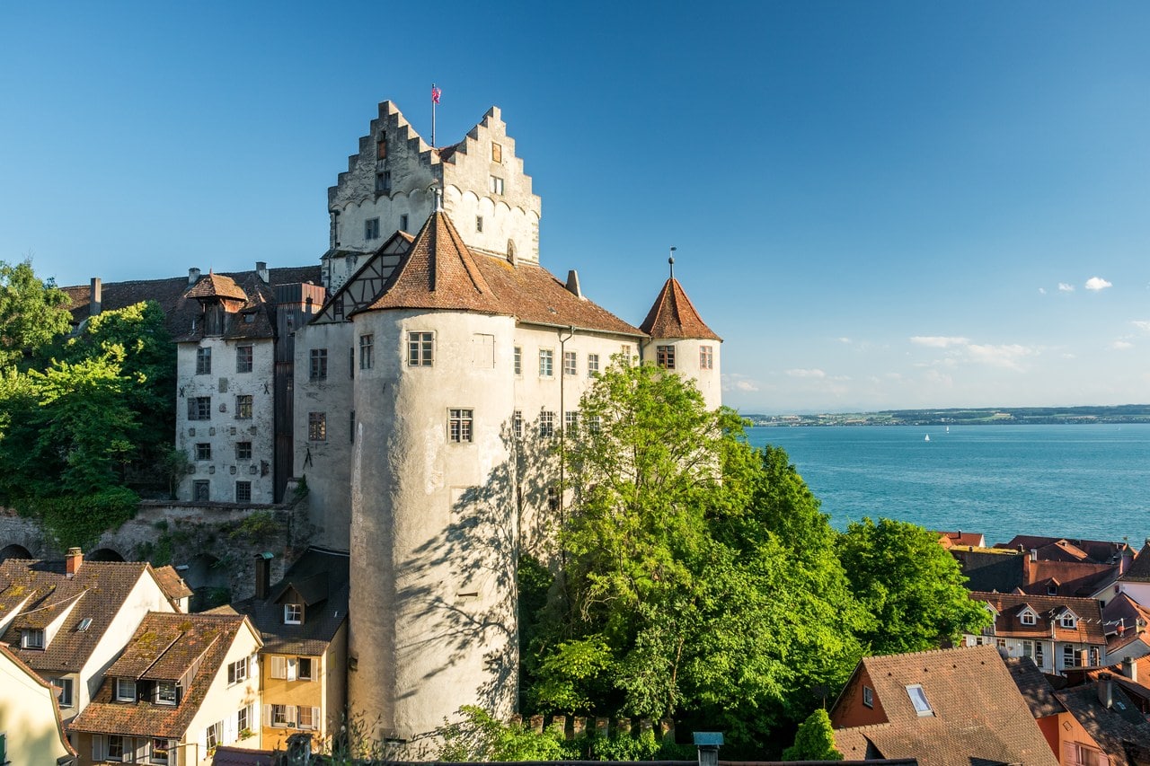 Burg Meersburg on a ridge above the tiled roofs of Meersburg old town, with a whitewashed block under a stepped gable and flagstaff, a tall round tower with a conical tiled roof in front of it, further castle ranges behind, and the blue water of Lake Constance to the right. Burg Meersburg on a ridge above the tiled roofs of Meersburg old town, with a whitewashed block under a stepped gable and flagstaff, a tall round tower with a conical tiled roof in front of it, further castle ranges behind, and the blue water of Lake Constance to the right.