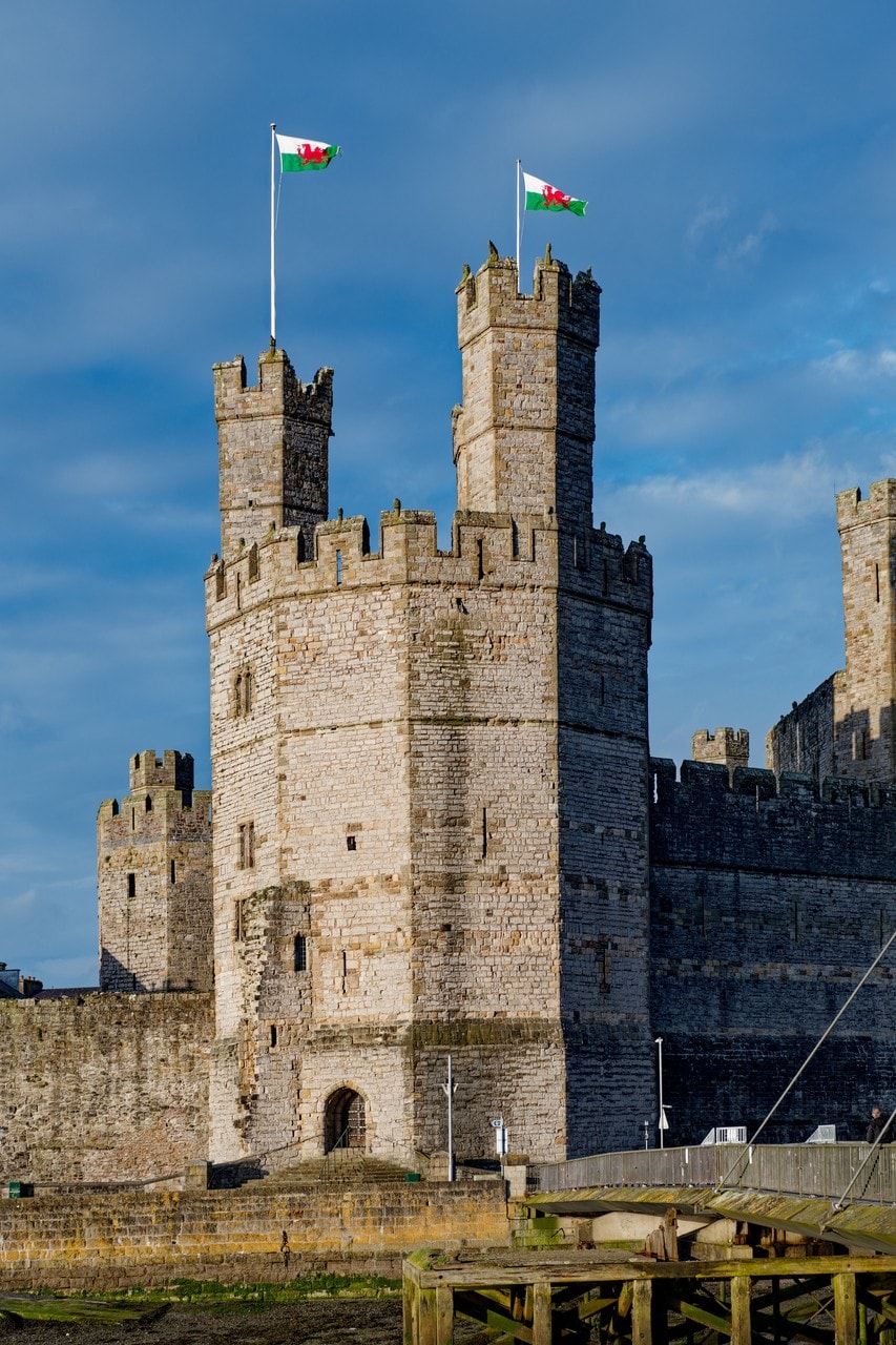 The polygonal Eagle Tower of Caernarfon Castle seen from below, its flat faces meeting at angles, with two small battlemented turrets rising from the battlements at the top, each flying a flag. The polygonal Eagle Tower of Caernarfon Castle seen from below, its flat faces meeting at angles, with two small battlemented turrets rising from the battlements at the top, each flying a flag.