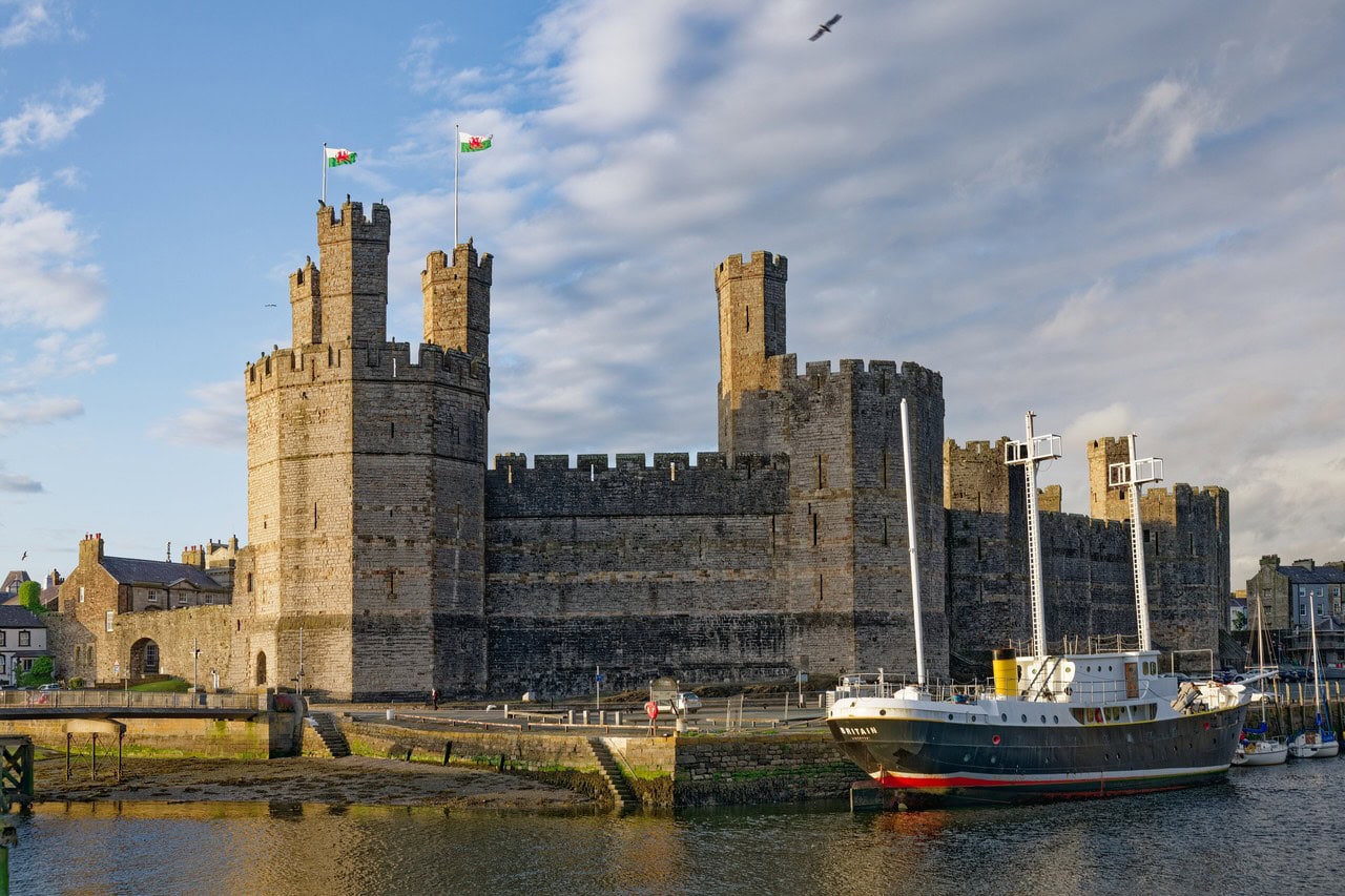 The south front of Caernarfon Castle seen across the water, with a large polygonal tower at the left carrying three small turrets, further polygonal towers ranged along the curtain wall, bands of darker stone in the masonry, and a moored ship in front of the wall. The south front of Caernarfon Castle seen across the water, with a large polygonal tower at the left carrying three small turrets, further polygonal towers ranged along the curtain wall, bands of darker stone in the masonry, and a moored ship in front of the wall.