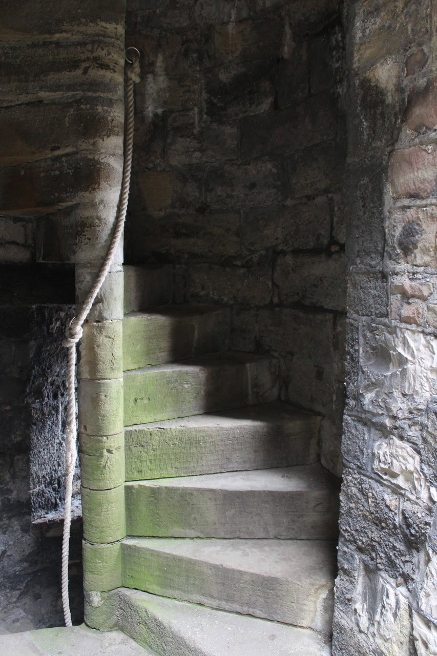 A stone spiral stair inside a castle tower, its wedge-shaped steps winding around a central newel formed by the stacked round inner ends of the steps, seen through a doorway with a rope handrail hanging beside it. A stone spiral stair inside a castle tower, its wedge-shaped steps winding around a central newel formed by the stacked round inner ends of the steps, seen through a doorway with a rope handrail hanging beside it.