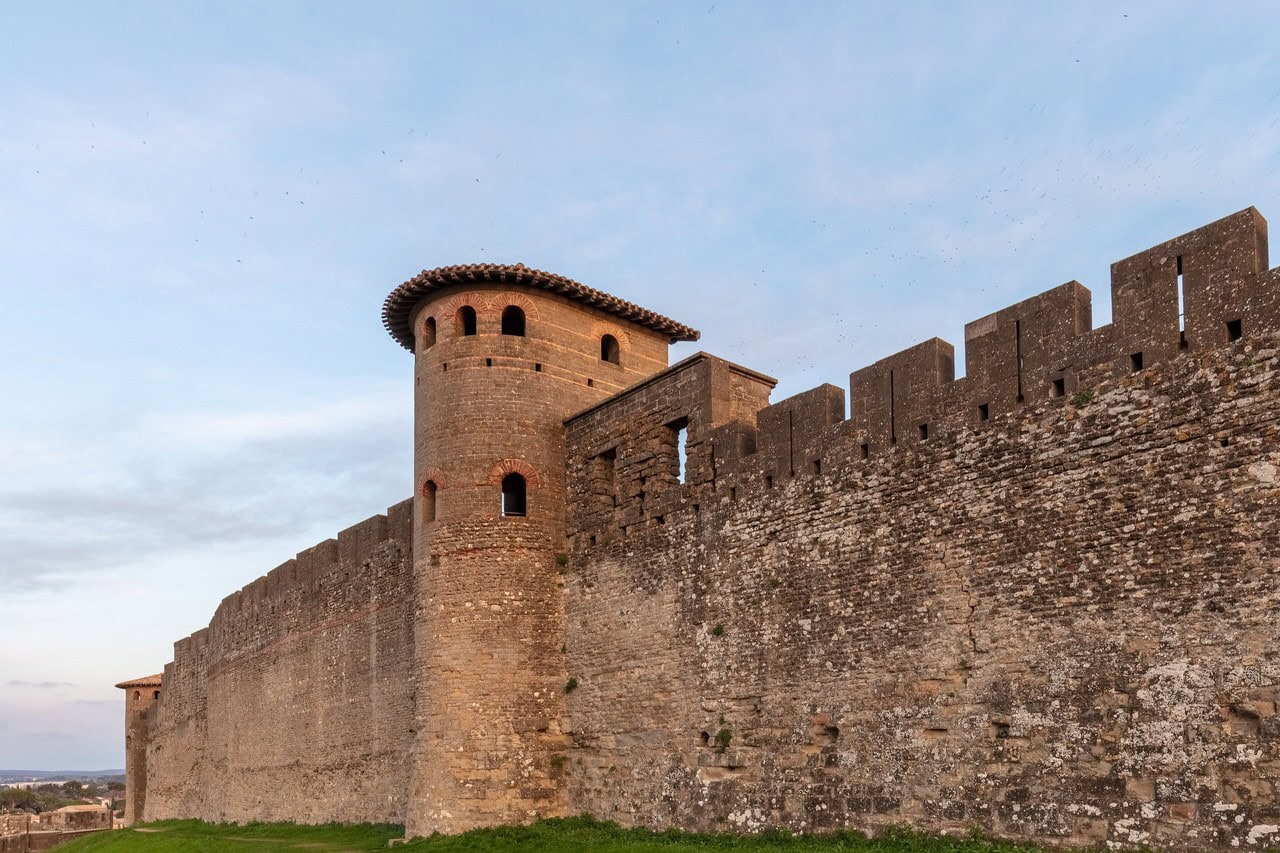 A half-round Gallo-Roman tower projecting from the stone curtain wall of Carcassonne, its curved face built of small coursed stone with bands of brick and pierced by narrow arched openings, under a tiled roof, with the crenellated curtain wall running away to the right. A half-round Gallo-Roman tower projecting from the stone curtain wall of Carcassonne, its curved face built of small coursed stone with bands of brick and pierced by narrow arched openings, under a tiled roof, with the crenellated curtain wall running away to the right.