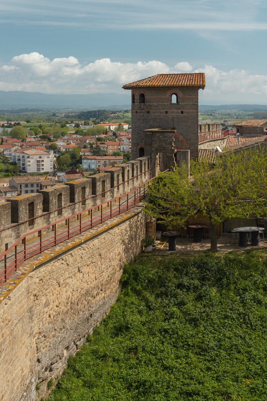 A tower of Carcassonne seen from a wall-walk, appearing rectangular under a hipped tile roof with brick bands running through its masonry, with a crenellated wall and a red steel handrail in the foreground and the lower town beyond. A tower of Carcassonne seen from a wall-walk, appearing rectangular under a hipped tile roof with brick bands running through its masonry, with a crenellated wall and a red steel handrail in the foreground and the lower town beyond.