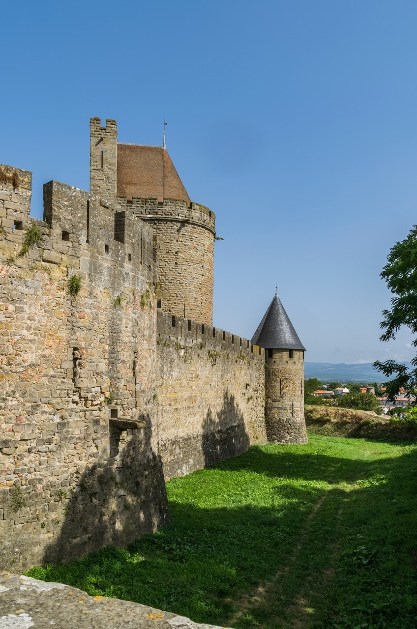A stretch of the walls of Carcassonne, with a large tower under a steep hipped tile roof at the left and a smaller round tower under a pointed slate cone standing further along the same wall, above a grass slope lying in the shadow of the rampart. A stretch of the walls of Carcassonne, with a large tower under a steep hipped tile roof at the left and a smaller round tower under a pointed slate cone standing further along the same wall, above a grass slope lying in the shadow of the rampart.