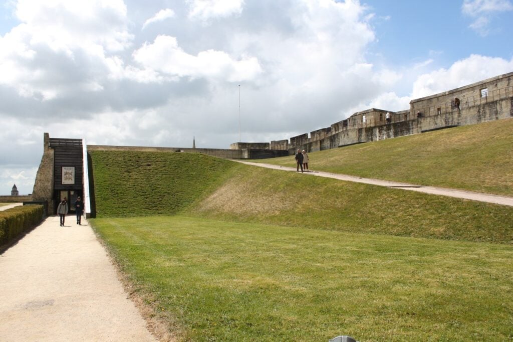 Château de Caen 5 A black timber entrance structure set into a grassed earthwork rampart at the Château de Caen, with the ashlar curtain wall and its wall walk beyond.