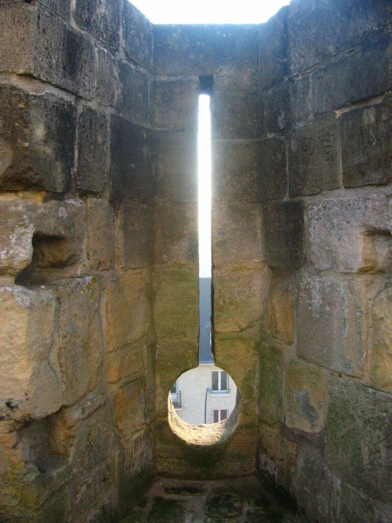 Château de Caen 10 The inside of a wall embrasure at the Château de Caen, a tall narrow loop ending in a circular opening, with splayed ashlar jambs on either side.