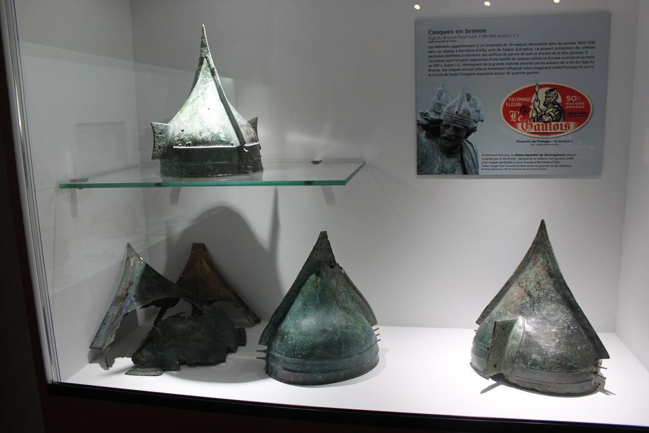 A lit museum case in the Musée de Normandie holding conical bronze helmets of the late Bronze Age, green with corrosion, with an interpretation panel behind them. A lit museum case in the Musée de Normandie holding conical bronze helmets of the late Bronze Age, green with corrosion, with an interpretation panel behind them.