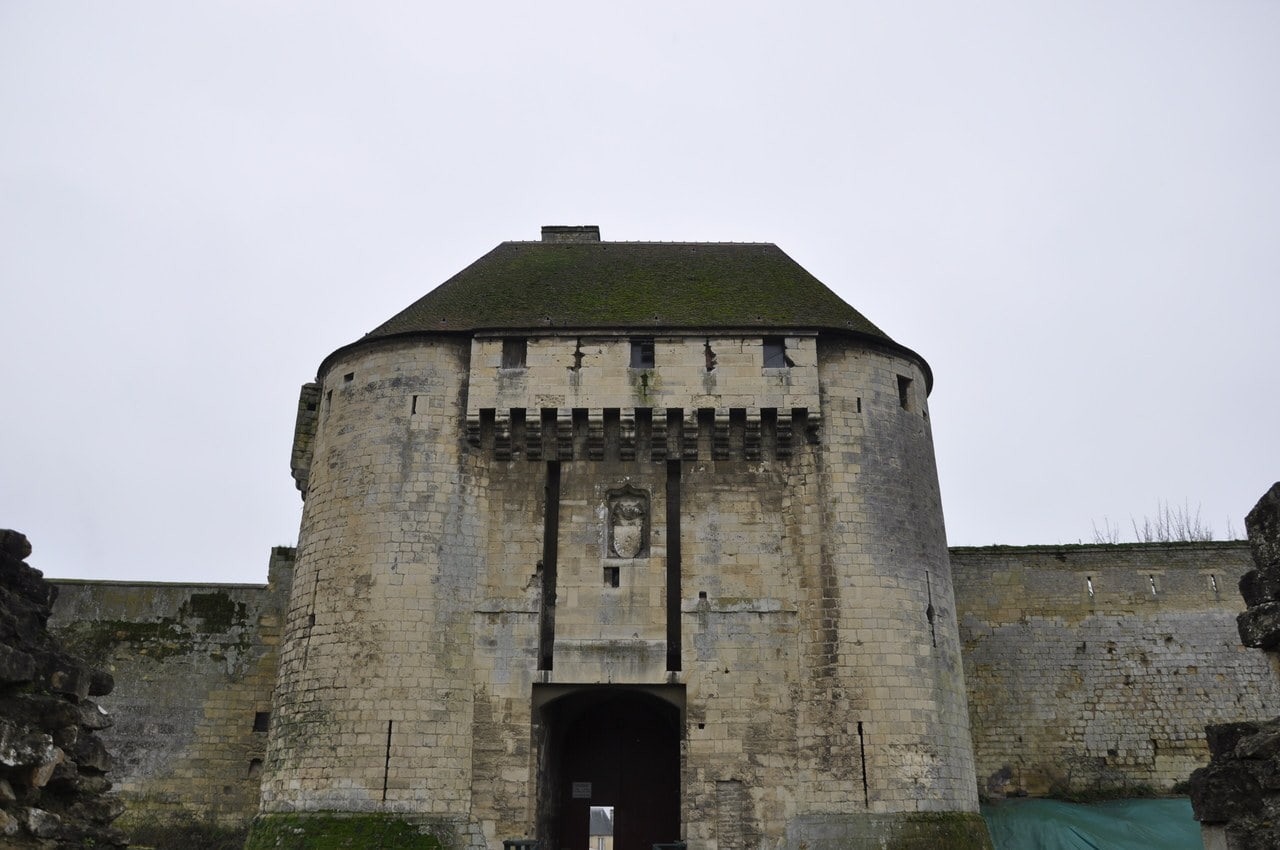 The rounded gate tower of the Porte des Champs at the Château de Caen, with a tiled hipped roof, a timber hoarding carried on stone corbels, an armorial panel above the arch, and the gate passage below. The rounded gate tower of the Porte des Champs at the Château de Caen, with a tiled hipped roof, a timber hoarding carried on stone corbels, an armorial panel above the arch, and the gate passage below.