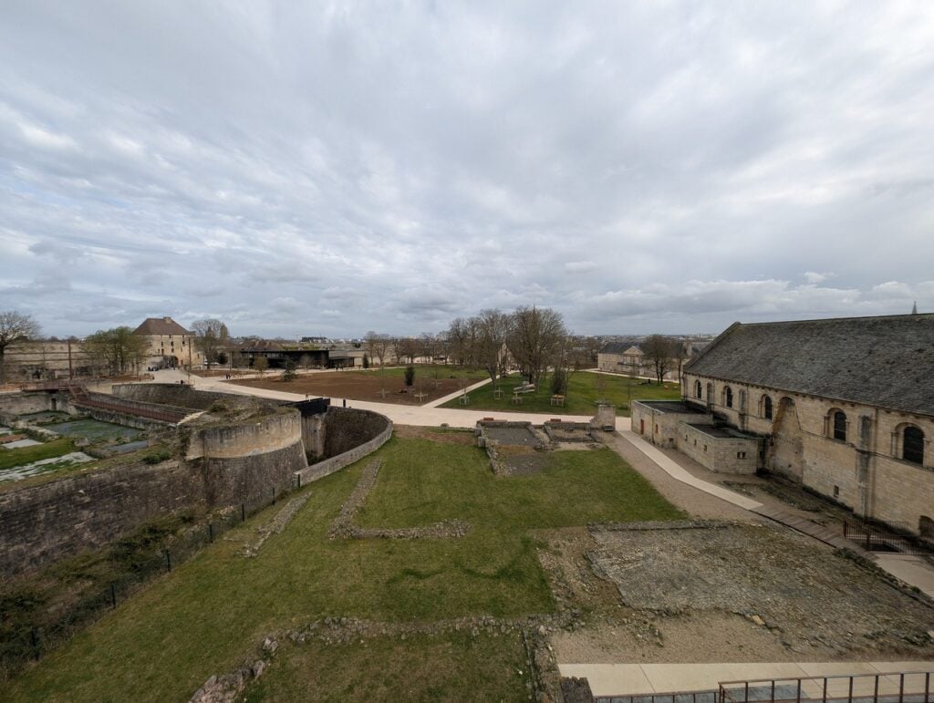 Château de Caen 9 Low excavated wall footings of the ducal palace laid out in mown grass at the Château de Caen, with the curved chemise wall of the donjon at left and the Salle de l'Échiquier at right.
