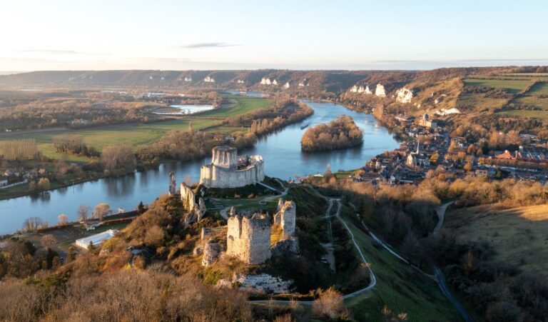 Home 7 Château Gaillard on its chalk spur at low sun, with the Seine looping around a wooded island and Le Petit-Andely on the bank below the castle.