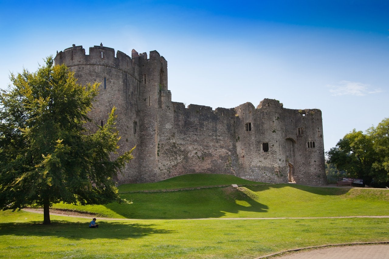 Chepstow Castle seen across mown grass from the Castle Dell, with a large drum tower at the left, a long crenellated curtain wall running away above a grassed ditch, and roofless ranges of domestic buildings standing behind the wall. Chepstow Castle seen across mown grass from the Castle Dell, with a large drum tower at the left, a long crenellated curtain wall running away above a grassed ditch, and roofless ranges of domestic buildings standing behind the wall.