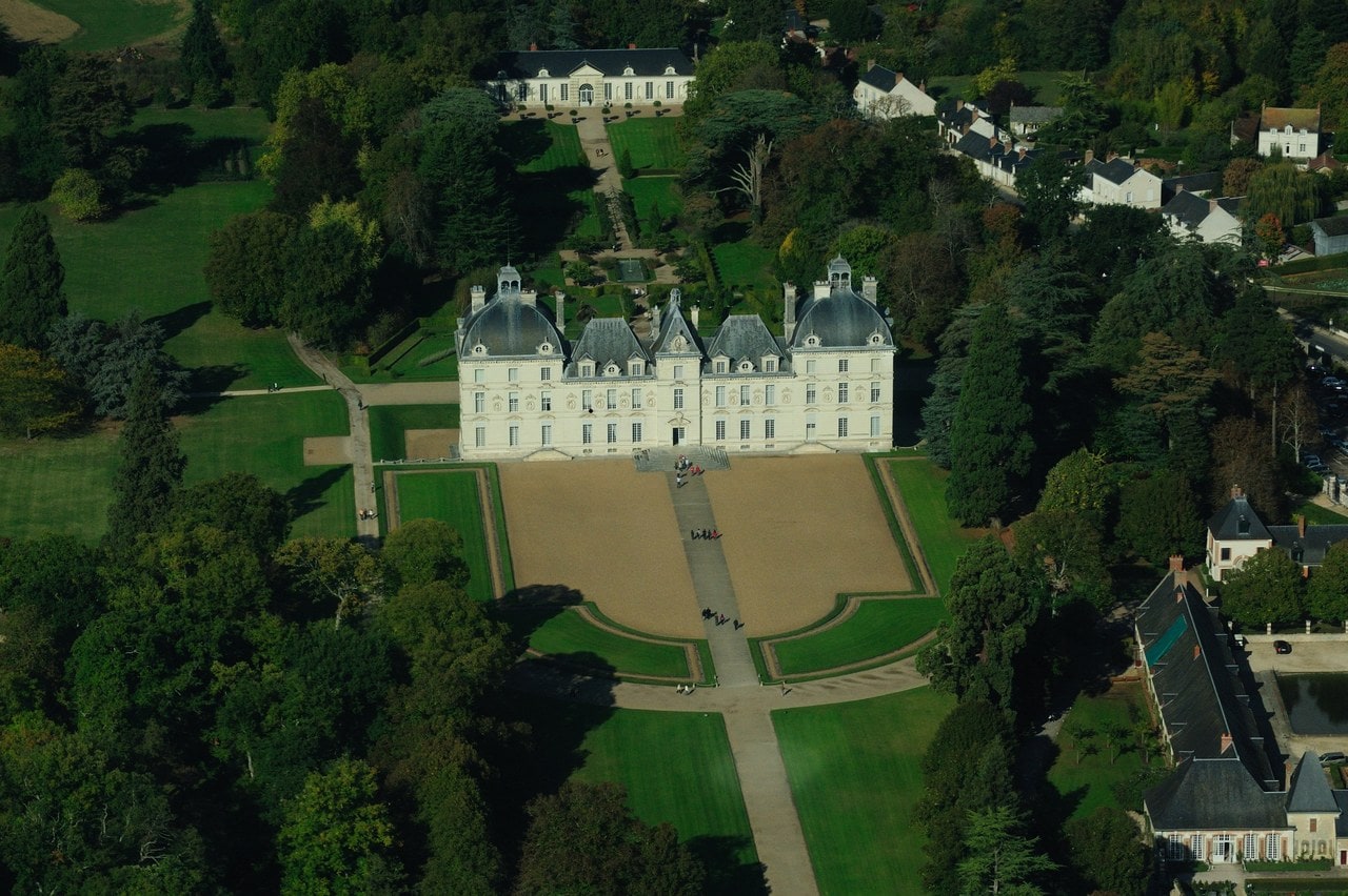 An aerial view of Chateau de Cheverny: a long white classical house with slate-domed end pavilions, a gravel forecourt and shaped lawns in front of it, a low pedimented garden pavilion at the head of the walk, and long slate-roofed service ranges to one side. An aerial view of Chateau de Cheverny: a long white classical house with slate-domed end pavilions, a gravel forecourt and shaped lawns in front of it, a low pedimented garden pavilion at the head of the walk, and long slate-roofed service ranges to one side.