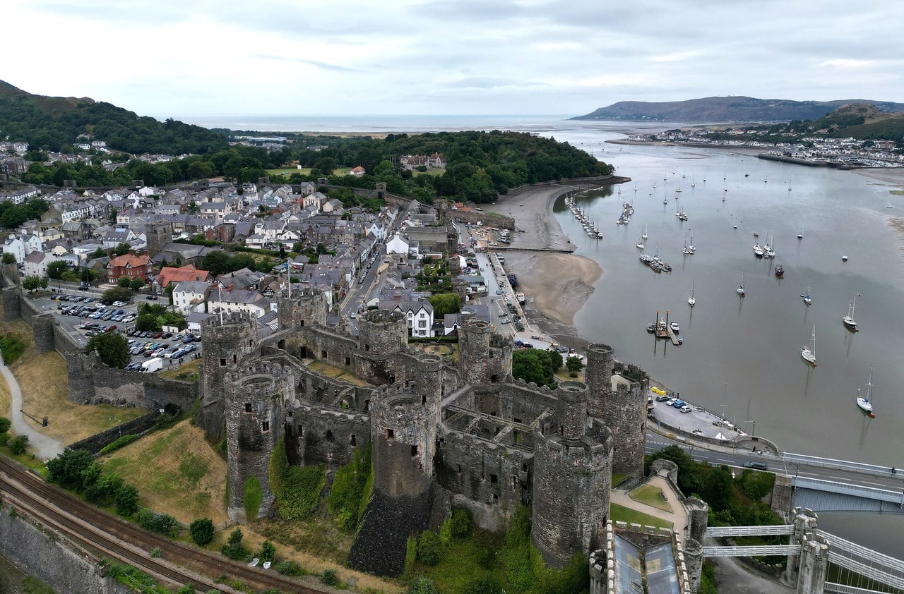 An aerial view of Conwy: the castle with its eight round towers on the waterfront in the foreground, the town of slate roofs and streets rising behind it, and a towered stone wall running around the town. An aerial view of Conwy: the castle with its eight round towers on the waterfront in the foreground, the town of slate roofs and streets rising behind it, and a towered stone wall running around the town.