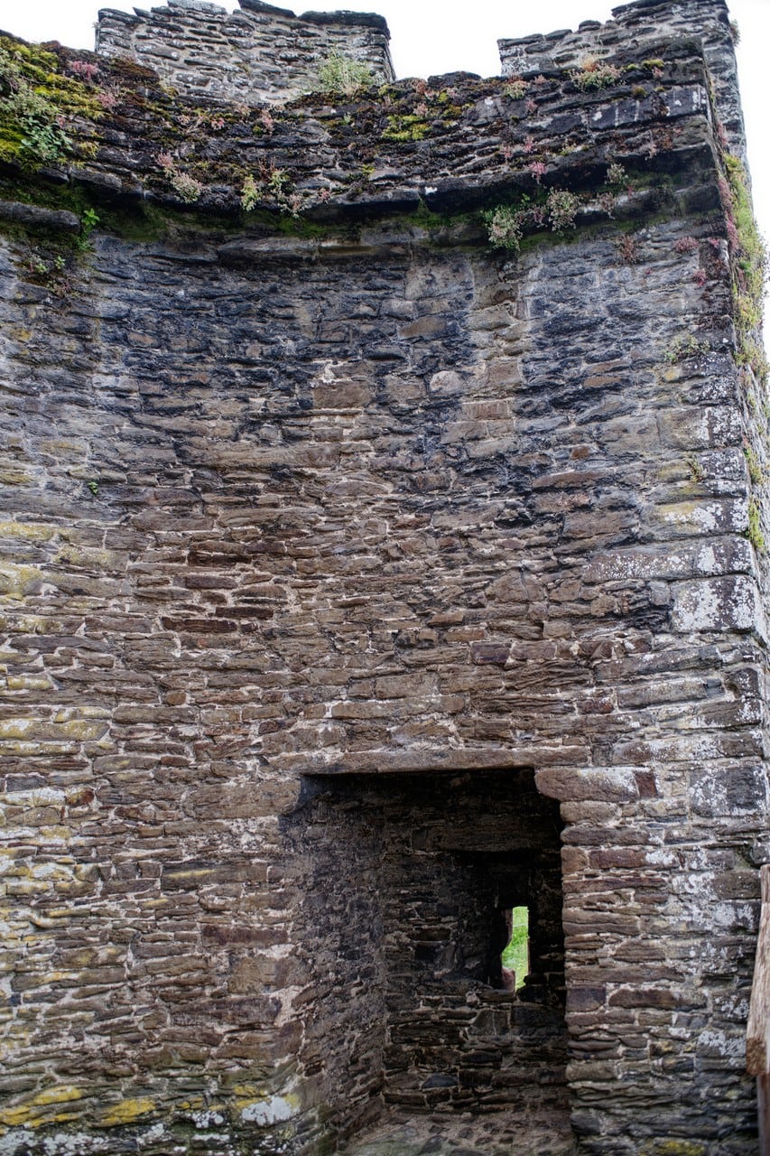 Looking into the open back of a tower on the Conwy town wall: a tall curved inner face of thin coursed rubble rising to battlements, with a broad square-headed recess at the bottom and daylight showing through a narrow slit inside it. Looking into the open back of a tower on the Conwy town wall: a tall curved inner face of thin coursed rubble rising to battlements, with a broad square-headed recess at the bottom and daylight showing through a narrow slit inside it.