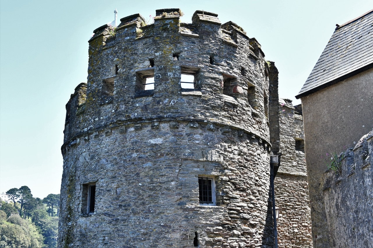 A broad round stone tower at Dartmouth Castle seen from below, with a ring of square-headed gun openings spaced around its upper stage, battlements above, an adjoining wall to the right and a slate roof beyond. A broad round stone tower at Dartmouth Castle seen from below, with a ring of square-headed gun openings spaced around its upper stage, battlements above, an adjoining wall to the right and a slate roof beyond.