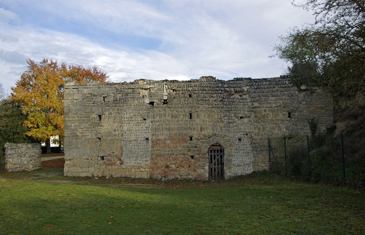 A roofless rectangular stone building at Doué-la-Fontaine standing in mown grass, built of coursed limestone in several visibly different patches, with rows of small square sockets in the walls, a barred iron grille over the doorway and a wire fence beyond. A roofless rectangular stone building at Doué-la-Fontaine standing in mown grass, built of coursed limestone in several visibly different patches, with rows of small square sockets in the walls, a barred iron grille over the doorway and a wire fence beyond.