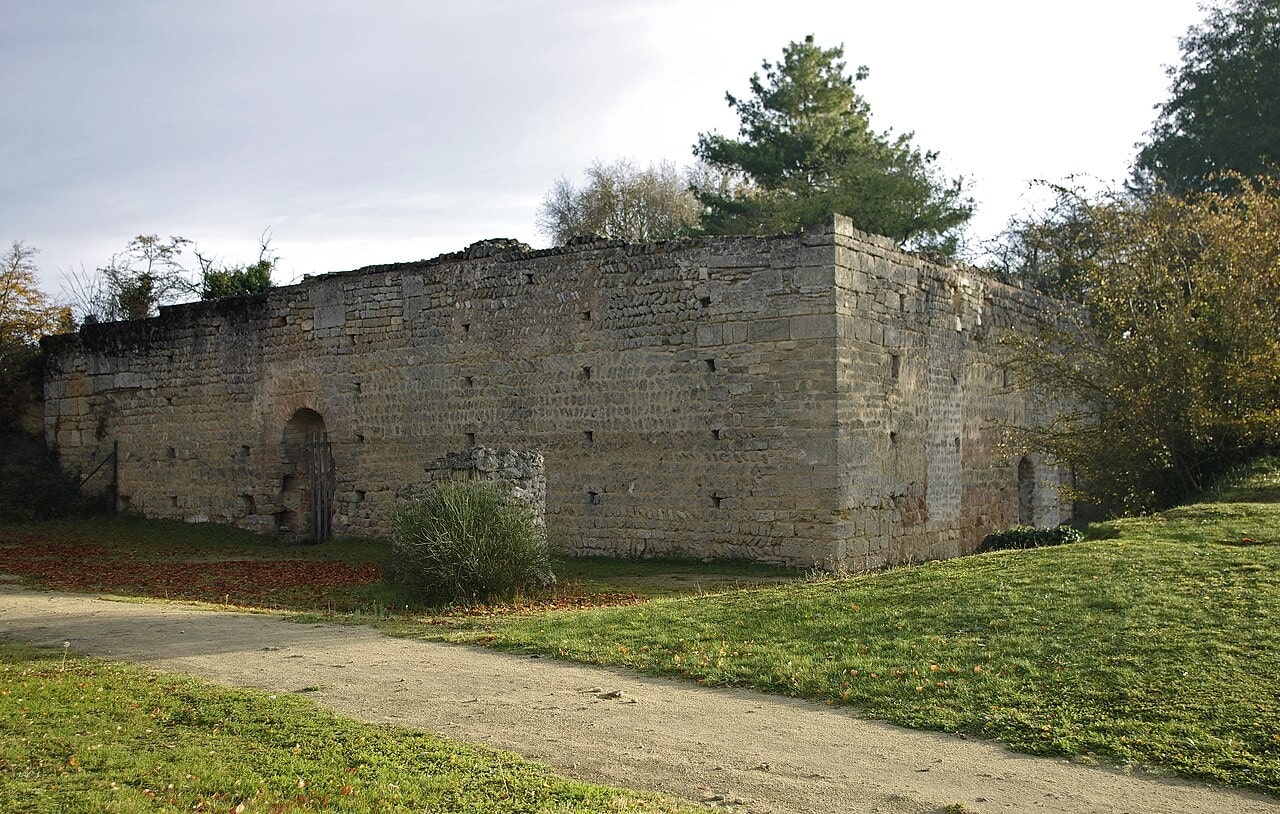 A roofless rectangular stone building seen across its corner so that a long side and the end wall both show, with large squared quoins at the angle, banded coursed rubble pierced by rows of small square sockets, a round-headed doorway closed by an iron gate, and a concrete path across the grass in front. A roofless rectangular stone building seen across its corner so that a long side and the end wall both show, with large squared quoins at the angle, banded coursed rubble pierced by rows of small square sockets, a round-headed doorway closed by an iron gate, and a concrete path across the grass in front.