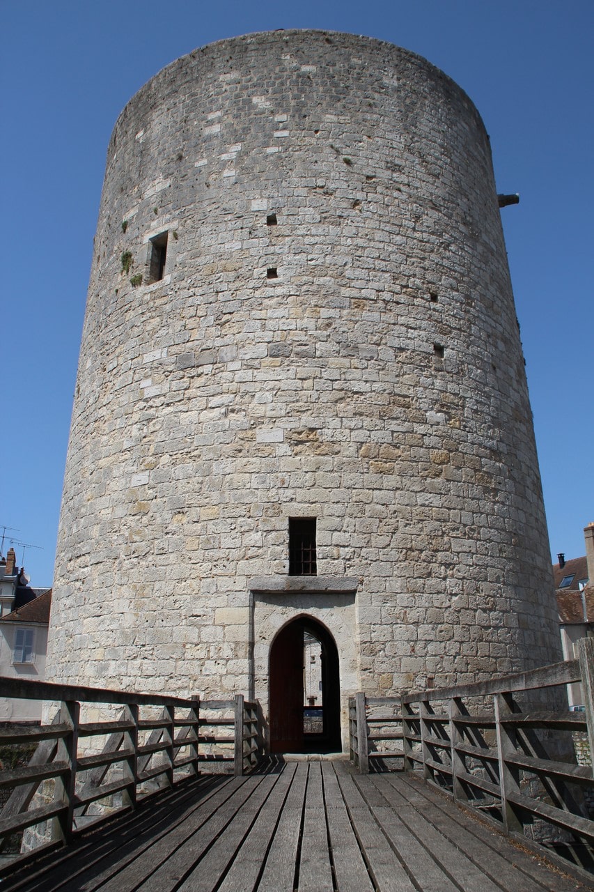 The round stone donjon of the château of Dourdan seen head on, a plain cylinder widening to a battered base, with a pointed-arched doorway at ground level reached by a modern timber bridge with wooden handrails.