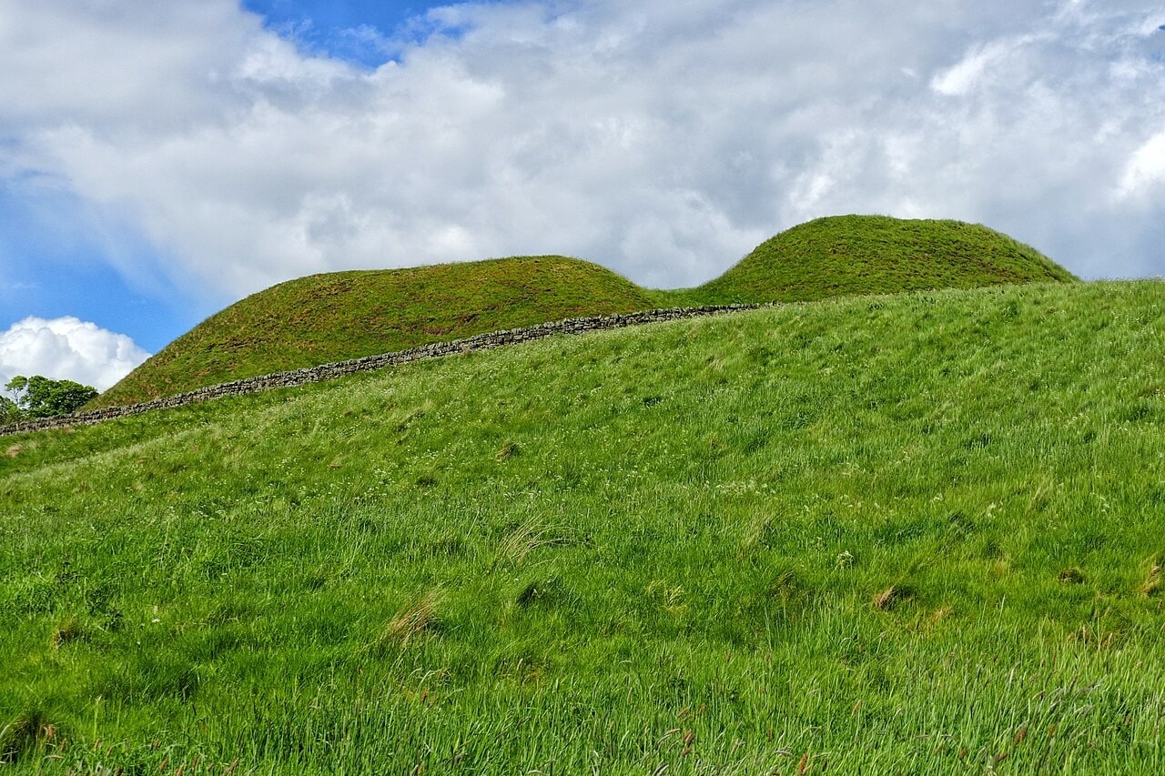 Two grassed earthworks on a skyline seen from below across a steep pasture: a long flat-topped bank on the left and a higher rounded mound on the right, with a drystone wall running across the slope between them and the viewer. Two grassed earthworks on a skyline seen from below across a steep pasture: a long flat-topped bank on the left and a higher rounded mound on the right, with a drystone wall running across the slope between them and the viewer.