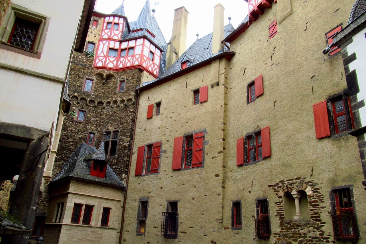 The inner courtyard of Burg Eltz looking up: a pale rendered wall on the left, a rubble-stone block in the middle with a red and white half-timbered oriel projecting near the roof and a slate-roofed turret below it, and on the right a tall ochre-rendered front with rows of red-shuttered windows. The inner courtyard of Burg Eltz looking up: a pale rendered wall on the left, a rubble-stone block in the middle with a red and white half-timbered oriel projecting near the roof and a slate-roofed turret below it, and on the right a tall ochre-rendered front with rows of red-shuttered windows.