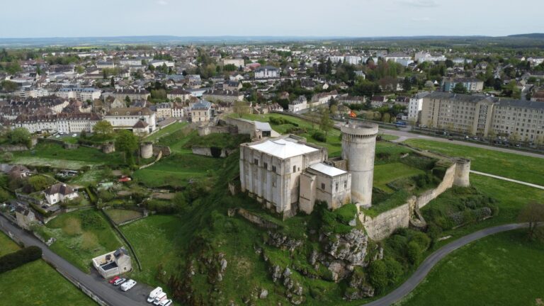 Home 5 Oblique aerial view of Chateau de Falaise on its rock, the great tower and a lower attached block under pale modern tent roofs, a round tower beside them, the walled enclosure spreading around them and the town of Falaise and open country beyond.