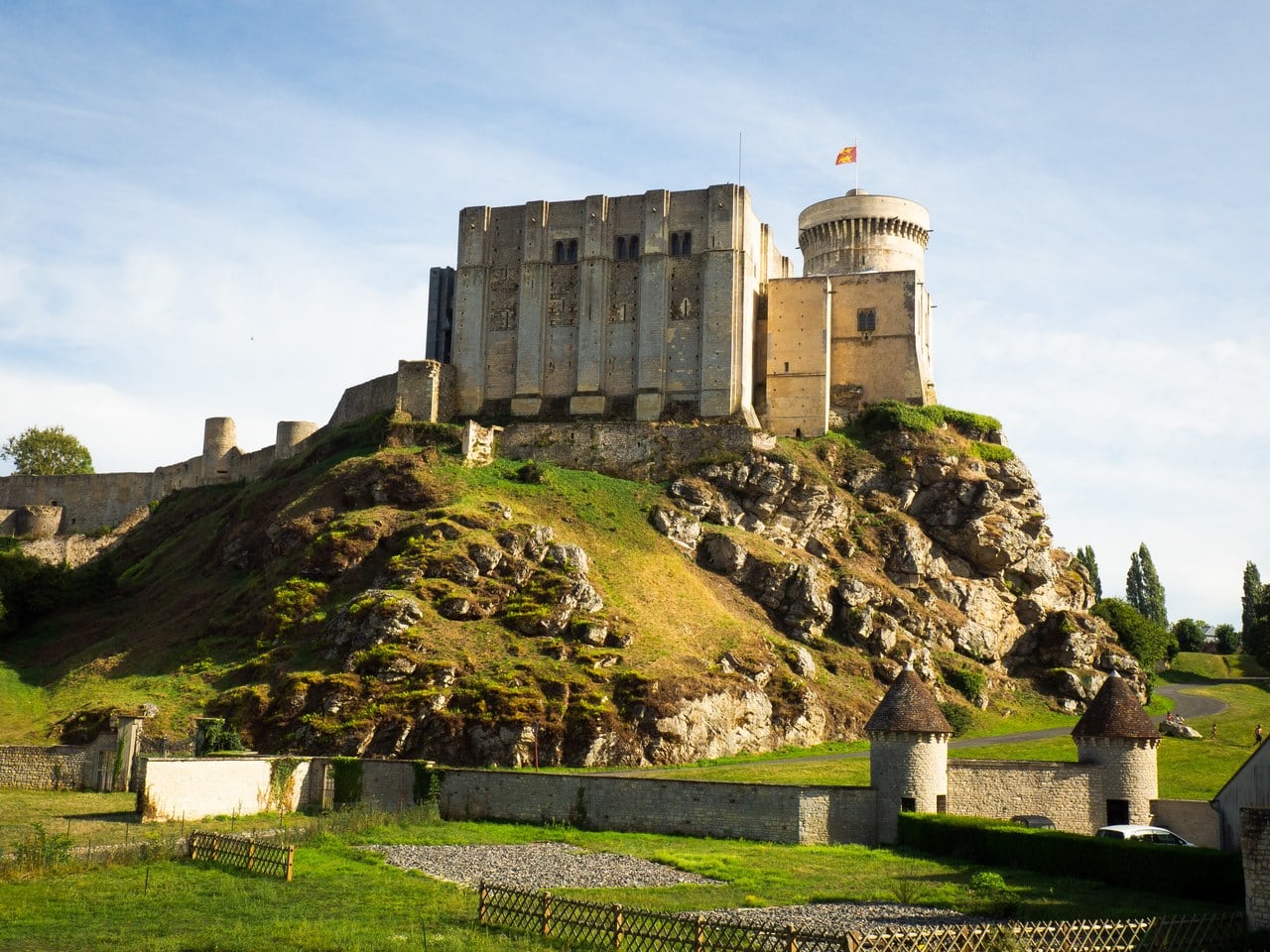Chateau de Falaise on a steep grassy knoll with bare limestone breaking through it, the great tower with shallow pilaster buttresses standing on the outcrop and the round Tour Talbot to its right, a low garden wall with two small conical-roofed turrets in the foreground. Chateau de Falaise on a steep grassy knoll with bare limestone breaking through it, the great tower with shallow pilaster buttresses standing on the outcrop and the round Tour Talbot to its right, a low garden wall with two small conical-roofed turrets in the foreground.