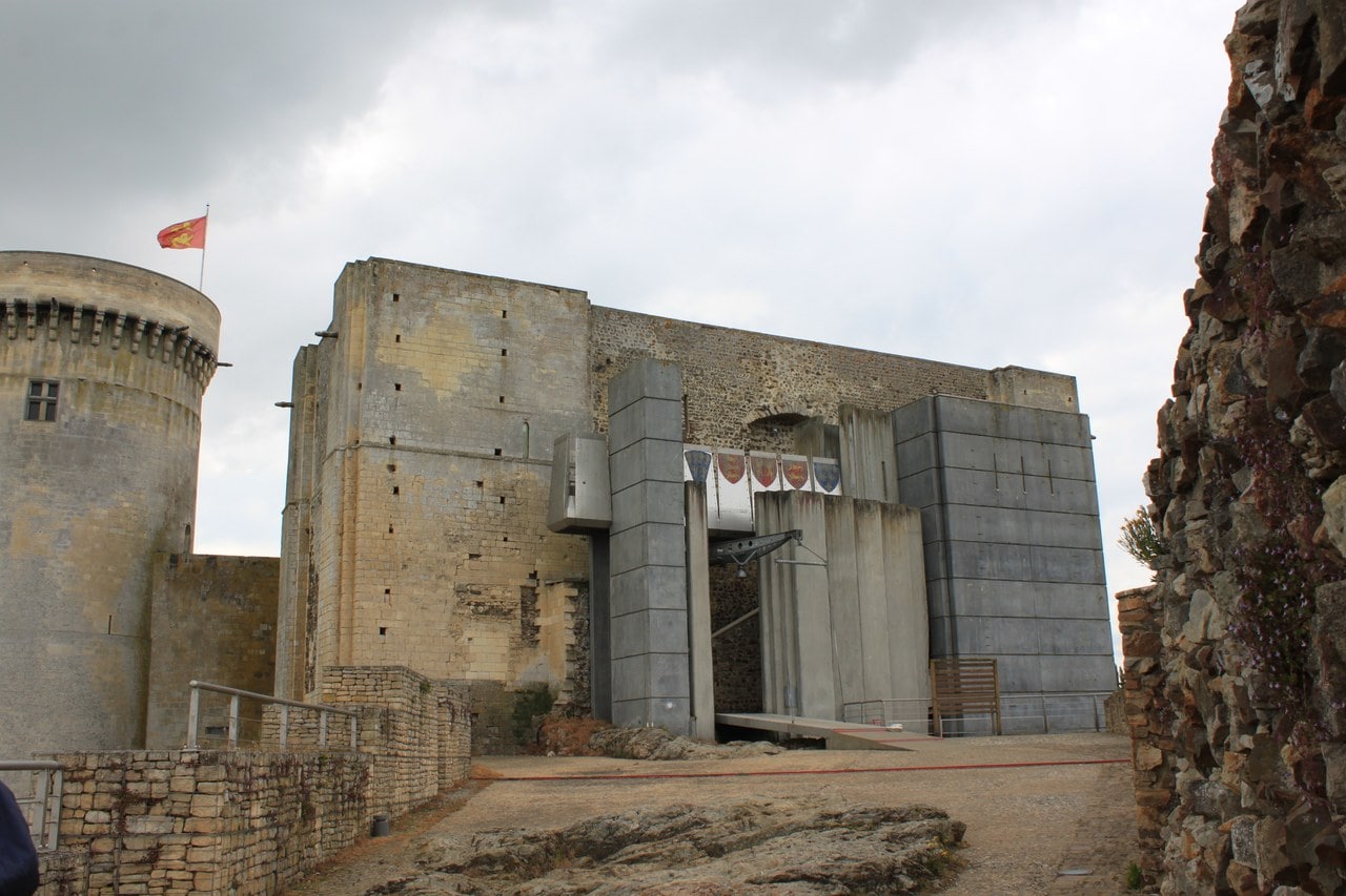 The entrance front of the great tower at Chateau de Falaise, older limestone walling at the left meeting tall dark gray concrete piers and a flat concrete face at the right, heraldic banners hung in the recessed entrance, a metal jib above a ramp, and the round Tour Talbot with a flag at the far left. The entrance front of the great tower at Chateau de Falaise, older limestone walling at the left meeting tall dark gray concrete piers and a flat concrete face at the right, heraldic banners hung in the recessed entrance, a metal jib above a ramp, and the round Tour Talbot with a flag at the far left.