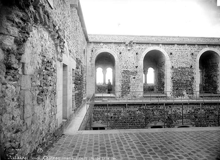 Château de Falaise 4 An early photograph of the roofless great hall at Chateau de Falaise, a row of round-headed windows in the far wall above a paved floor, with a timber rail and a single standing figure.