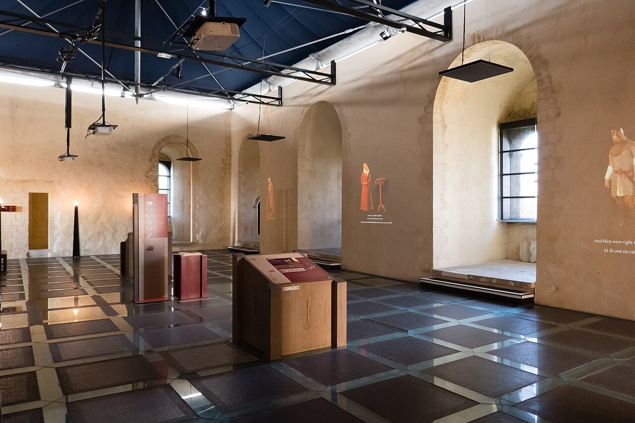 A large hall inside the great tower at Chateau de Falaise, pale rendered walls with round-headed arches and a deep window embrasure, a black steel truss ceiling hung with projectors and lights, projected figures and bilingual text on the walls, and a floor of large dark reflective slabs set in a pale grid. A large hall inside the great tower at Chateau de Falaise, pale rendered walls with round-headed arches and a deep window embrasure, a black steel truss ceiling hung with projectors and lights, projected figures and bilingual text on the walls, and a floor of large dark reflective slabs set in a pale grid.