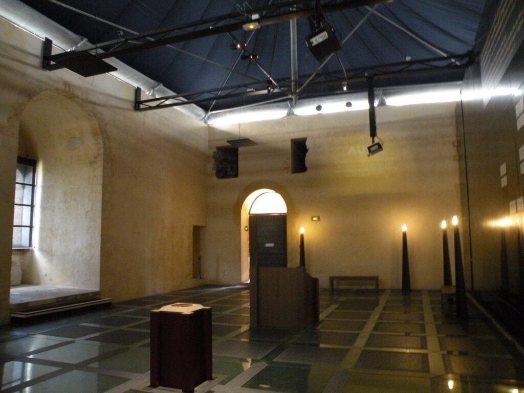 Château de Falaise 3 A large rendered hall at Chateau de Falaise with a round-headed arched opening, roofed by a black steel truss hung with theatrical lights, over a checkered modern floor with dark conical display objects standing on it.