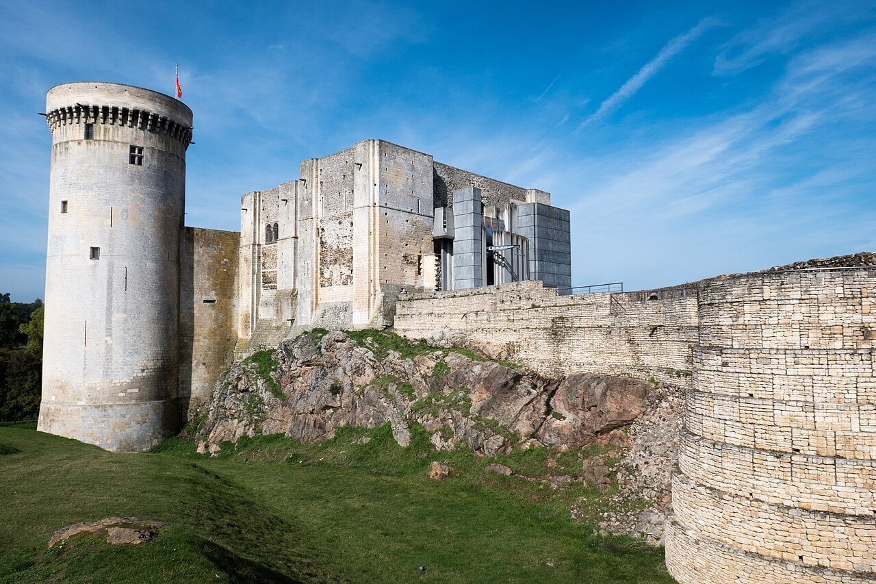 The keep block at Chateau de Falaise, rectangular masses of pale limestone with flat pilaster buttresses standing hard against one another, the round Tour Talbot with its corbelled parapet at the left, a gray concrete structure at the far end, and a large curved tower of new coursed ashlar in the right foreground. The keep block at Chateau de Falaise, rectangular masses of pale limestone with flat pilaster buttresses standing hard against one another, the round Tour Talbot with its corbelled parapet at the left, a gray concrete structure at the far end, and a large curved tower of new coursed ashlar in the right foreground.
