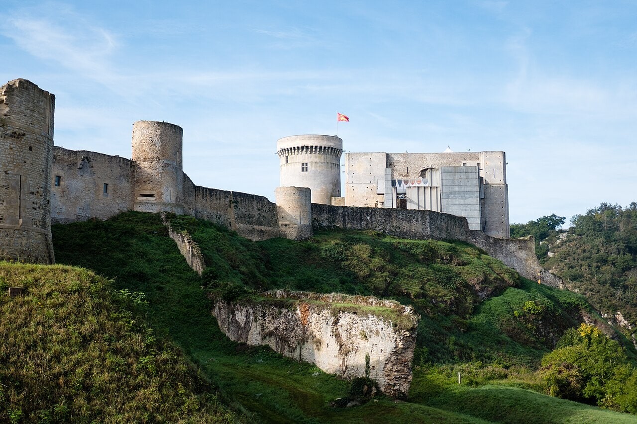 Chateau de Falaise seen from below on the north side, a curtain wall with mural towers running along the crest of the rock, the round Tour Talbot with a corbelled parapet, and the great tower behind it with a gray concrete structure hung with banners against its face. Chateau de Falaise seen from below on the north side, a curtain wall with mural towers running along the crest of the rock, the round Tour Talbot with a corbelled parapet, and the great tower behind it with a gray concrete structure hung with banners against its face.