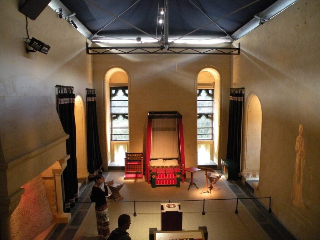 Château de Falaise 7 A staged chamber inside the small donjon at Chateau de Falaise, a red hung bed and wooden chests below tall arched windows, a carved figure at the right and visitors at the lower left.