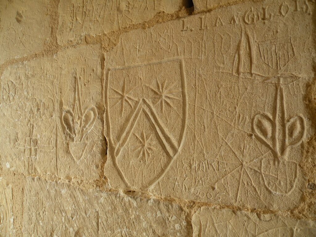 Château de Falaise 6 Incised graffiti on the pale ashlar wall of a chamber in the Tour Talbot at Chateau de Falaise, including shield shapes, a star, crosses and carved letters.