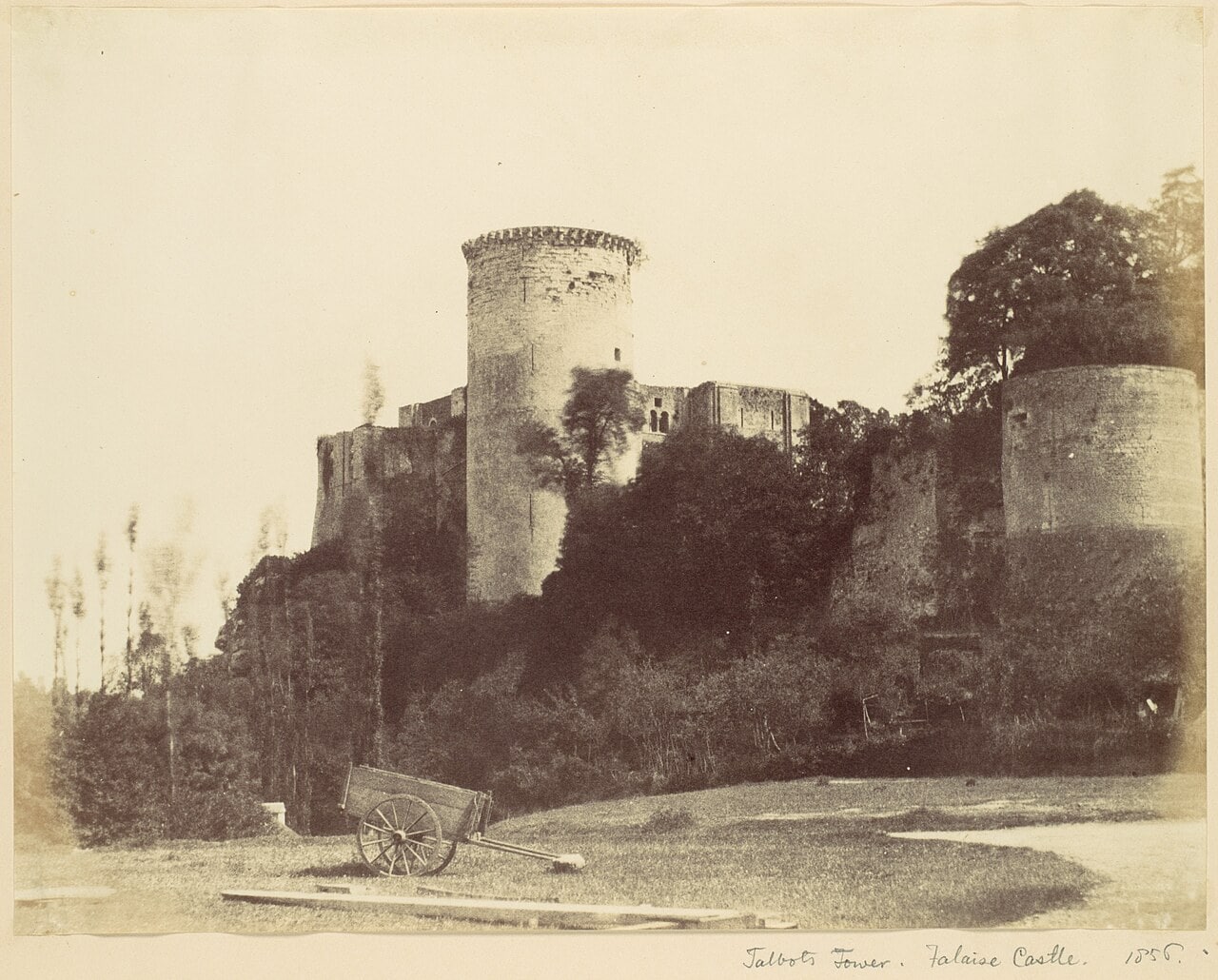 An 1856 photograph of the Tour Talbot at Chateau de Falaise, a tall cylindrical tower with a corbelled parapet and ivy over its flank, the roofless great tower behind it, a second large drum tower at the right, and a two-wheeled handcart standing on bare ground in the foreground. An 1856 photograph of the Tour Talbot at Chateau de Falaise, a tall cylindrical tower with a corbelled parapet and ivy over its flank, the roofless great tower behind it, a second large drum tower at the right, and a two-wheeled handcart standing on bare ground in the foreground.
