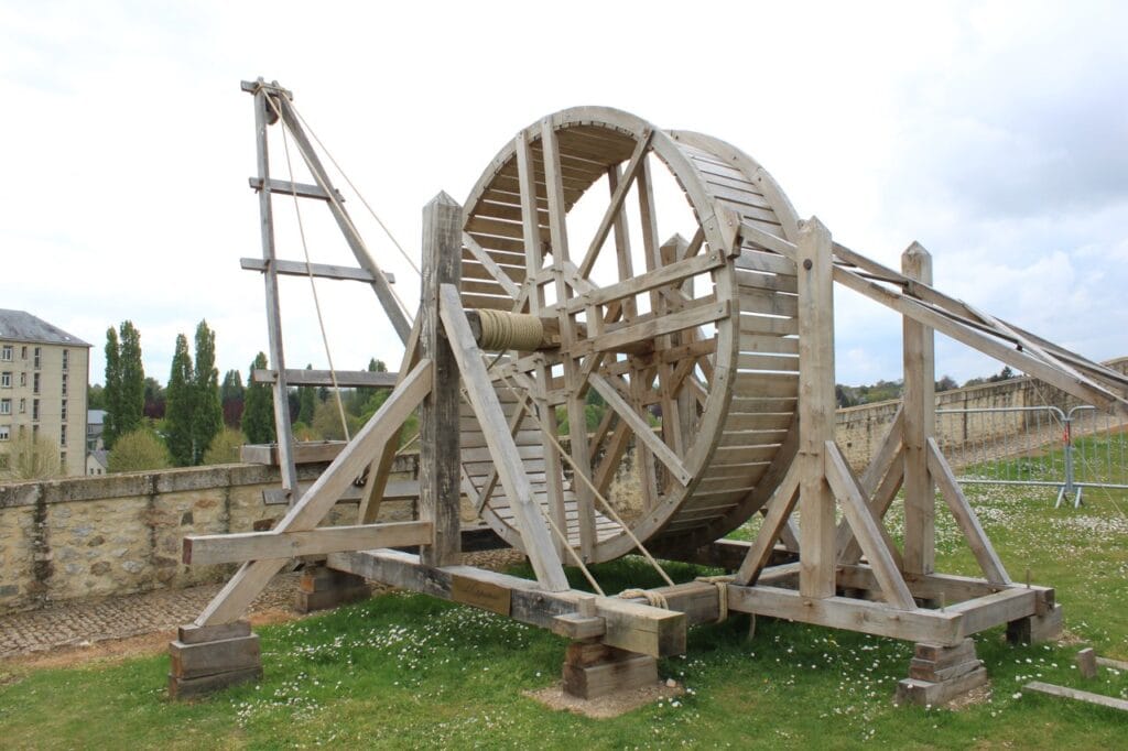 Château de Falaise 8 A large timber treadwheel crane reconstructed on the grass at Chateau de Falaise, its rope drum on the axle and a stone wall running behind it, with a second timber frame at the right.