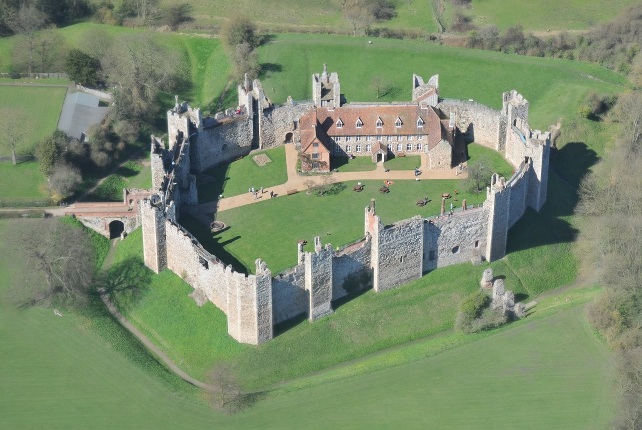 Aerial view of Framlingham Castle, a circuit of stone curtain wall with square projecting towers that stand open at the back, a grassy empty courtyard, and one brick range with dormer windows and tall chimneys inside the walls.