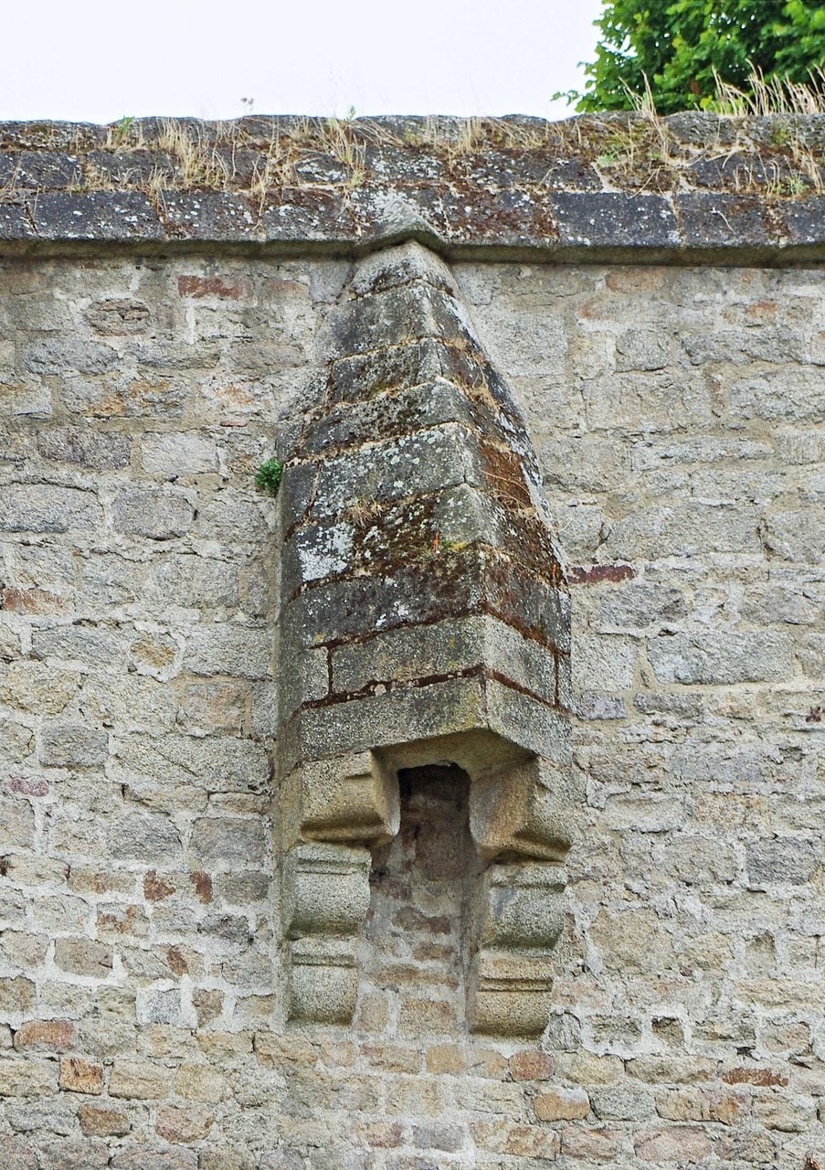 A tall stone shaft with a steeply gabled cap running down the outer face of a rubble wall, carried on moulded corbels, with an arched opening at its foot. A tall stone shaft with a steeply gabled cap running down the outer face of a rubble wall, carried on moulded corbels, with an arched opening at its foot.