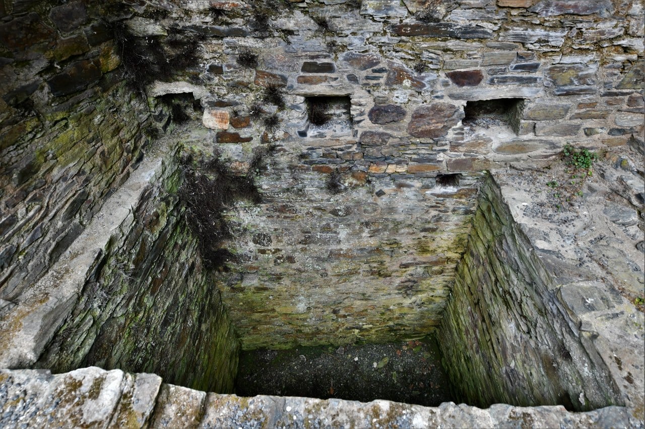 A deep rectangular pit lined with coursed stone, sunk below a ruined wall face, its lower courses green with moss and two small voids visible in the masonry above. A deep rectangular pit lined with coursed stone, sunk below a ruined wall face, its lower courses green with moss and two small voids visible in the masonry above.
