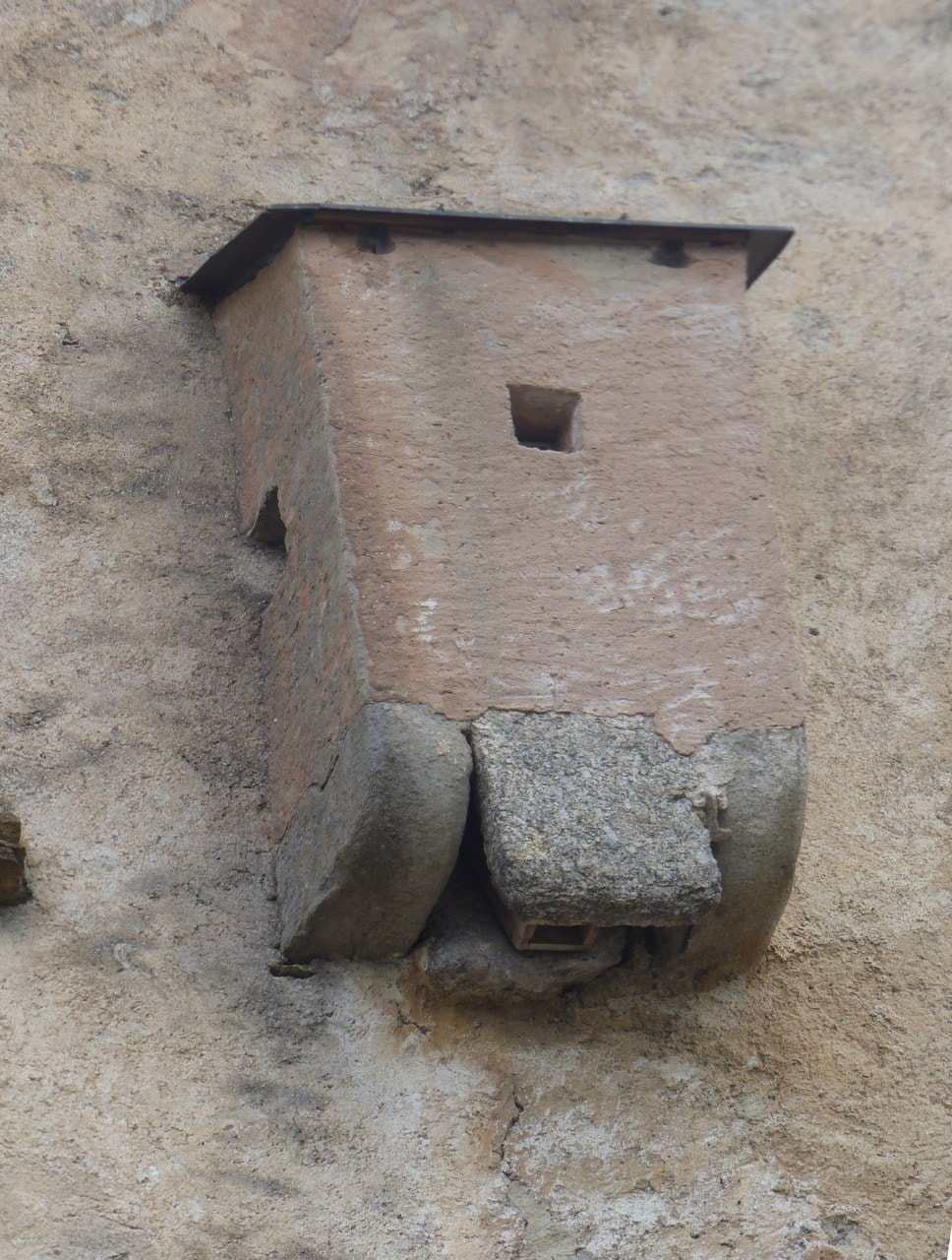 A rendered box corbelled out from a stone wall with a flat slab roof and a small square opening in its side; beneath it two shaped stone corbels carry the floor, and the discharge opening is visible under them. A rendered box corbelled out from a stone wall with a flat slab roof and a small square opening in its side; beneath it two shaped stone corbels carry the floor, and the discharge opening is visible under them.