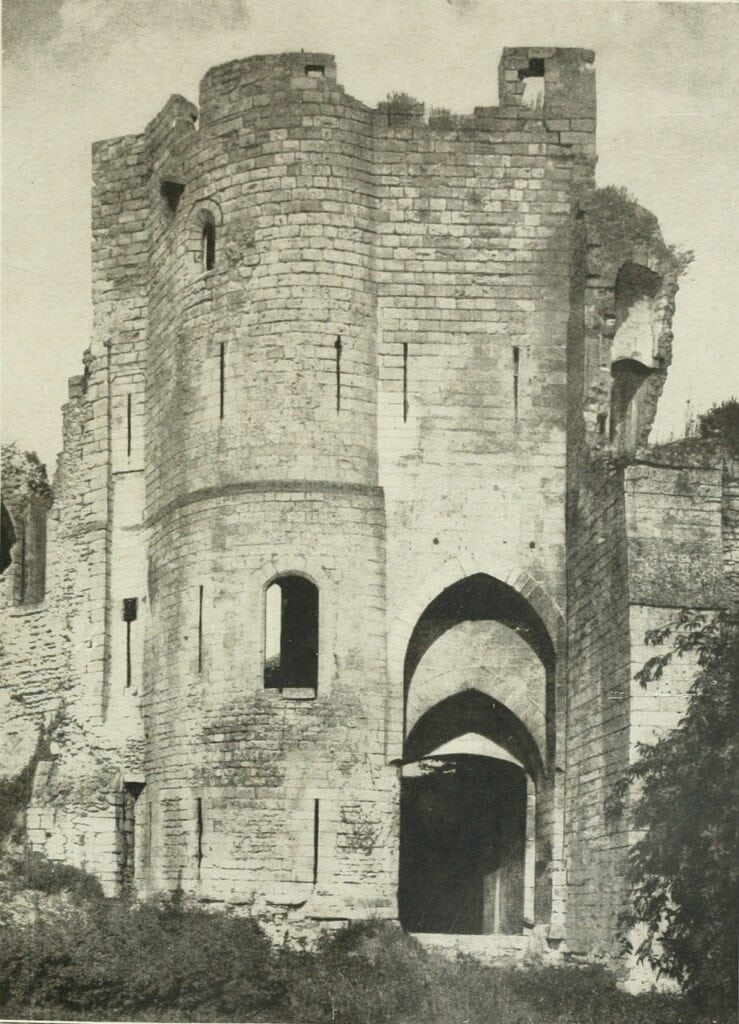 Château de Gisors 7 A black and white photograph of the outer gateway of Chateau de Gisors, a rounded flanking turret pierced with narrow arrow slits beside a two-order pointed archway, with rough grass running up to the threshold.