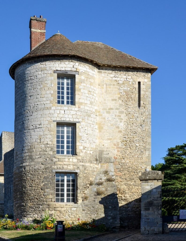 Château de Gisors 9 A round medieval mural tower at Chateau de Gisors under a conical tiled roof with a brick chimney, three tall multi-pane windows cut through its curved wall and a narrow arrow slit in the masonry to the right.