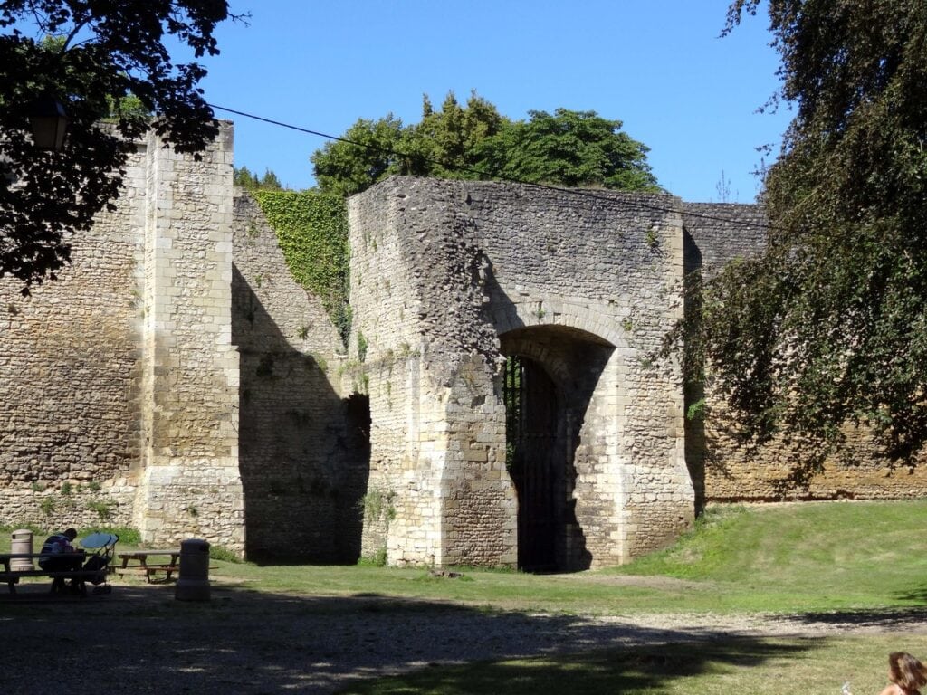 Château de Gisors 8 A ruined stone range inside the walls of Chateau de Gisors, a tall pointed archway barred with a metal grille at its center, ivy along the wall head and picnic tables on the grass in front.