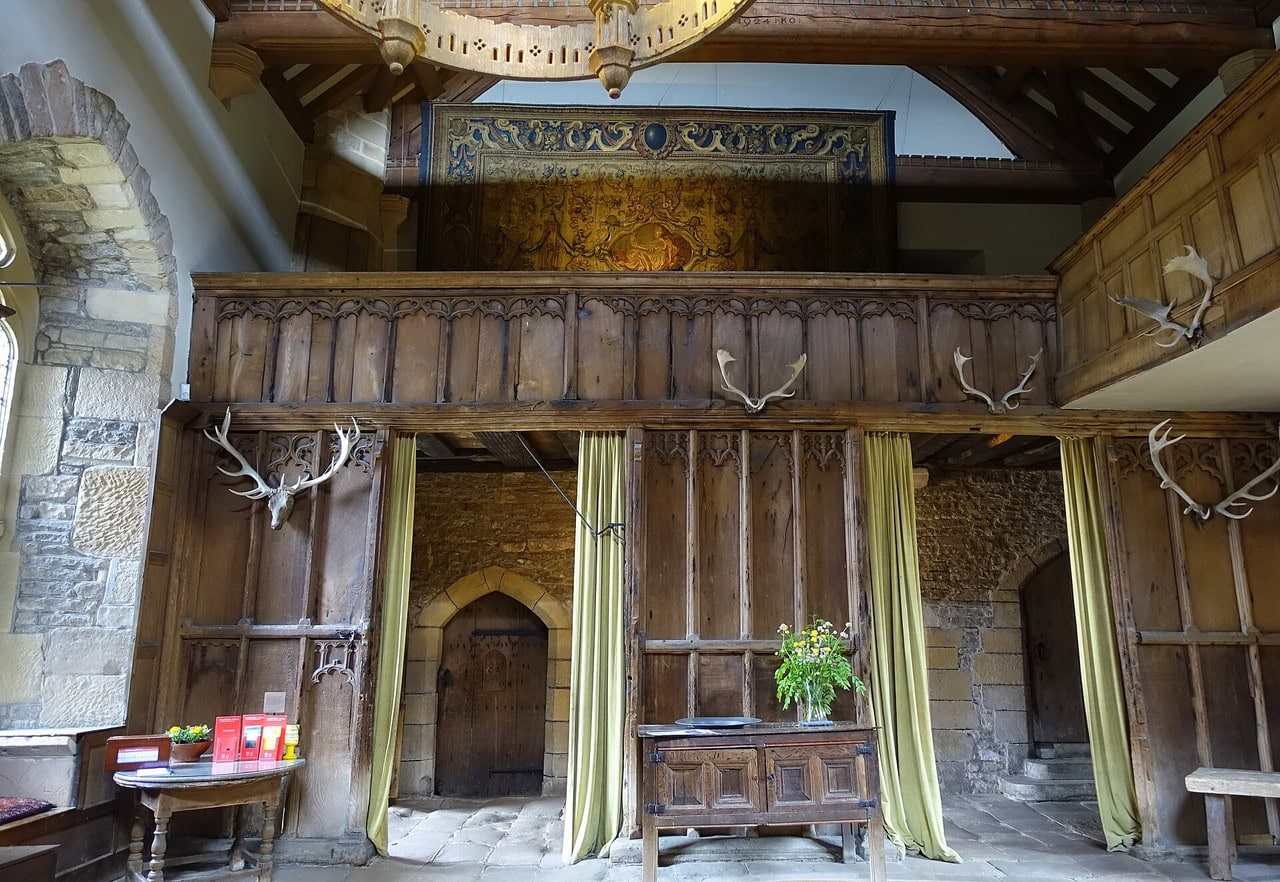 A carved timber screen closing the low end of a hall, with a traceried head, two openings hung with curtains, and a carved gallery above; a flagged passage and a stone stair are visible through the openings. A carved timber screen closing the low end of a hall, with a traceried head, two openings hung with curtains, and a carved gallery above; a flagged passage and a stone stair are visible through the openings.