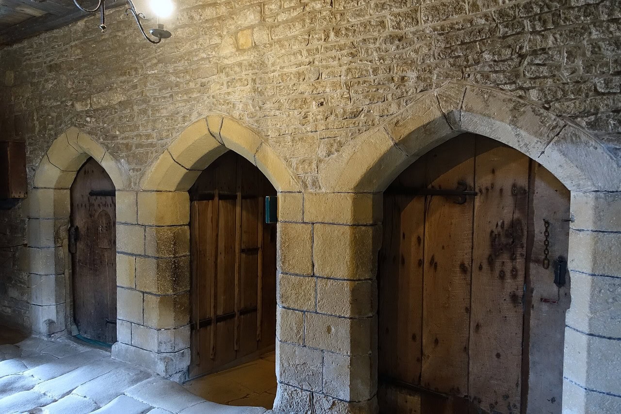 Three pointed stone doorways side by side off a flagged passage, each closed by a plank door with iron studs. Three pointed stone doorways side by side off a flagged passage, each closed by a plank door with iron studs.