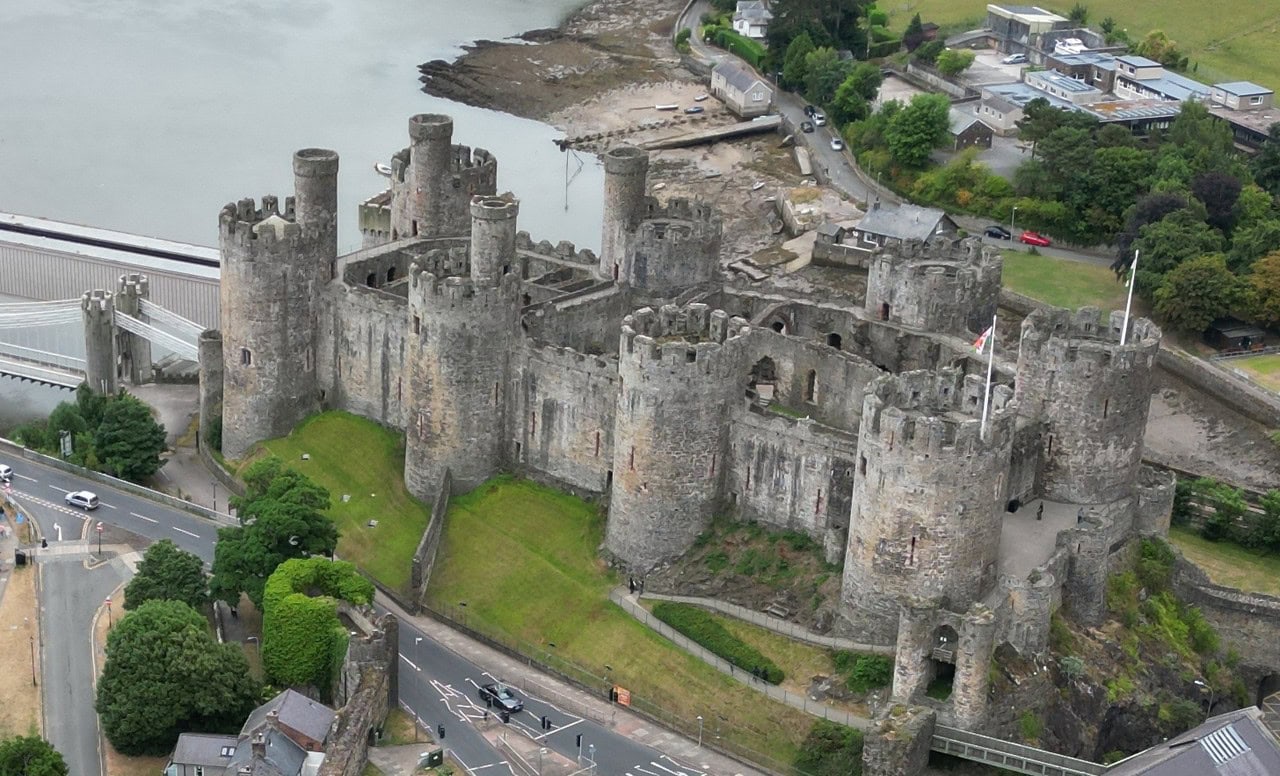 Castle Towers 1 Aerial view of Conwy Castle, a long walled enclosure whose curtain wall carries round towers projecting at regular intervals, several of them topped by slender turrets, with a grassy ditch and a road below and the estuary beyond.