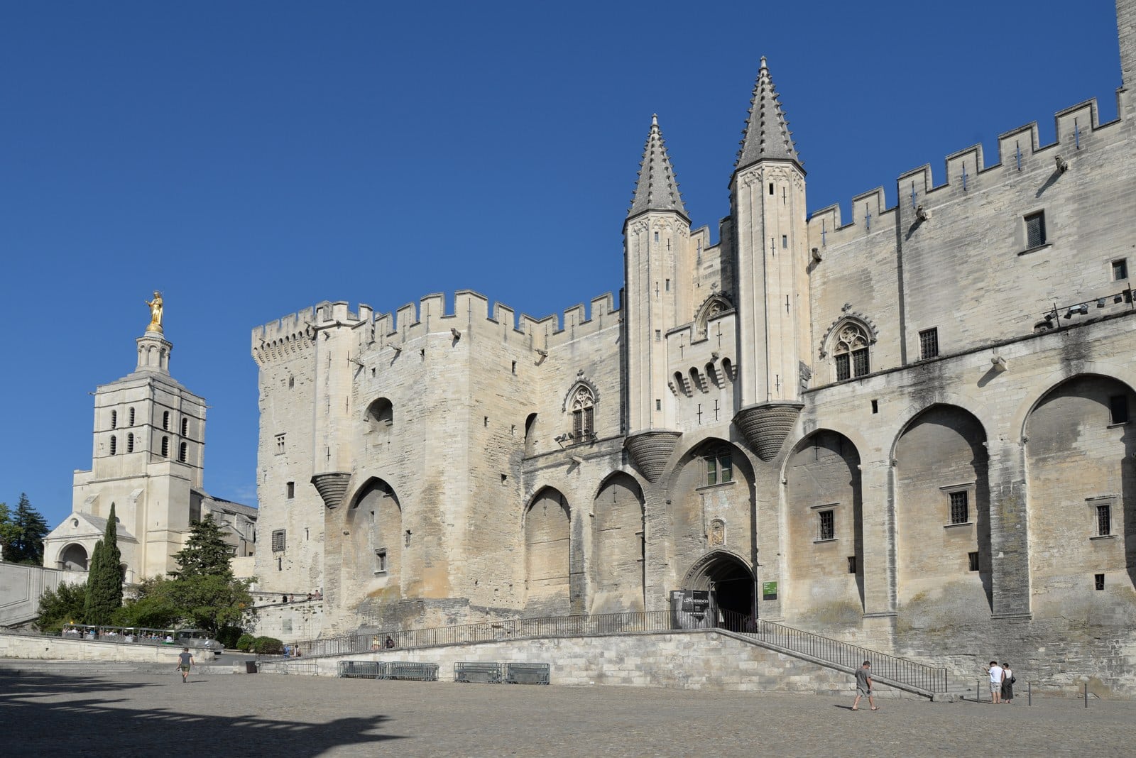Castle vs Palace vs Fortress 1 The west front of the Palais des Papes in Avignon: a crenellated stone wall with cross-shaped arrow loops and giant blind arches, and a gateway flanked by two pinnacled turrets, with the bell tower of Avignon Cathedral and its gilded Virgin at left.